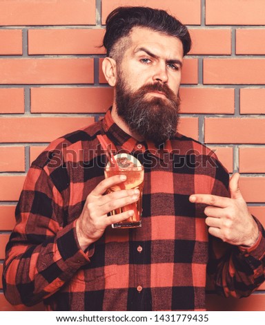 Man points at cocktail on brick wall background. Offer to drink concept. Barman with beard and inquiring face holds glass with cocktail straw. Hipster with stylish hair holds drink or cocktail.