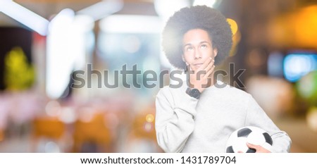 Young african american man with afro hair holding soccer football ball serious face thinking about question, very confused idea