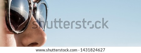 close-up image of woman's face in sunglasses reflecting rays of sun, copy space, blue sky background, panoramic shot