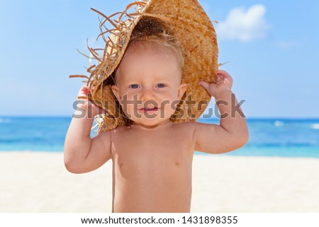 On sunny white sand beach muddy happy baby boy holds hat and has fun before swimming in sea waves. Active children lifestyle, summer vacation travel with kids on tropical family resort