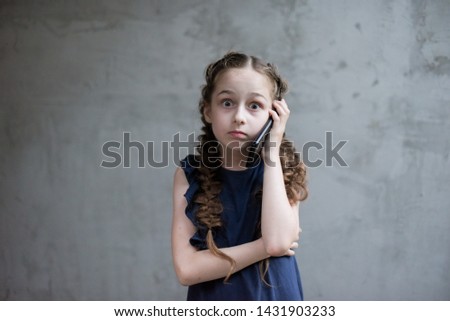 Beautiful little girl calling on mobile phone. Girl 8-9 years old emotionally talking on the phone at the gray wall. Beautiful little schoolgirl. Modern schoolchildren. School.