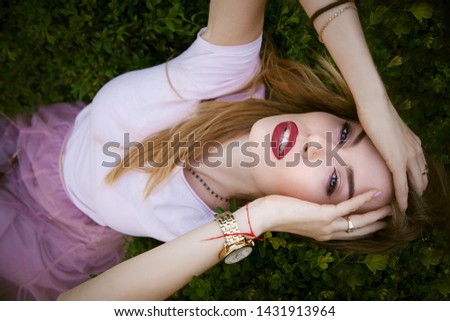 A luxurious young woman with long hair, red lips and a white T-shirt, tulle skirt sits with her head thrown back and rests her head on a green leafy bush, touching her head with her hands. Portrait.