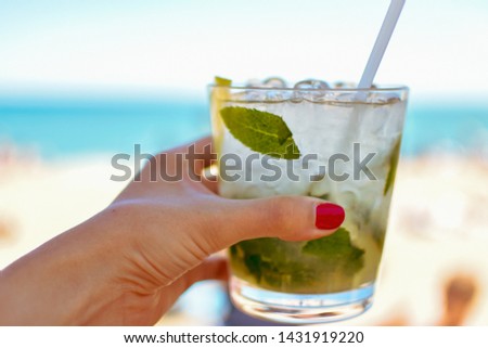 Woman's hand with red nail polish holding glass of fresh mojito cocktail with ice, mint, lime and lemons. Beach backdrop. Enhanced blue and green colors. Barcelona, Spain -Image