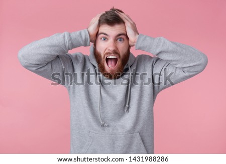 Portrait of young screaming shocked red bearded man in gray hoodie, looks evil and displeased , holds head, his favorite team lost. Stands over pink background and with wide open mouth.