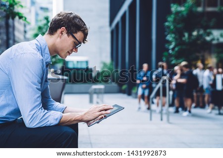 Side view of young successful man in spectacles for vision protection searching contact number for making online video conference via modern touch pad, concept of market digitalisation on tablet Royalty-Free Stock Photo #1431992873