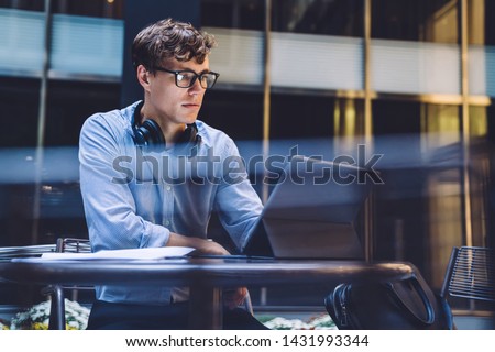 Young man skilled office worker using touch pad during break at job while sitting at cafe terrace, intelligent male proud ceo in classic eyeglasses reading incoming email with financial report Royalty-Free Stock Photo #1431993344