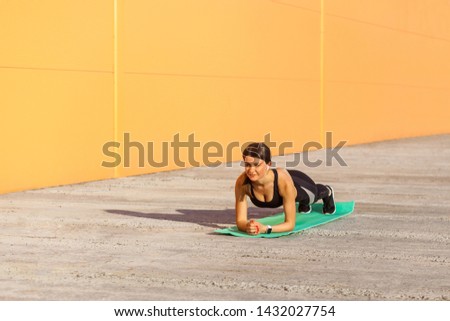 Muscular sporty strong beautiful young athletic woman in black sportwear standing on perfect plank position on elbows on green mat. Outdoor, orange wall background, sport and healthy concept