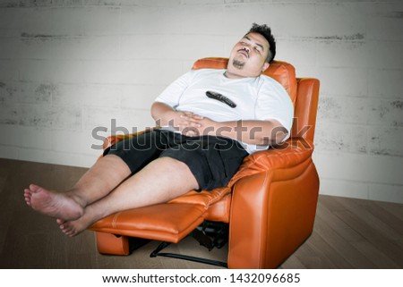 Picture of a young obese man falling asleep on the sofa while watching television in the living room