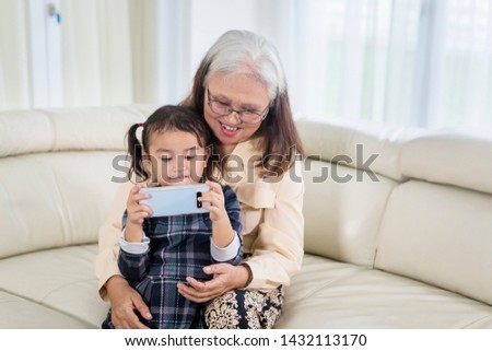 Picture of cute little girl using a mobile phone with her grandmother at home while sitting on the sofa