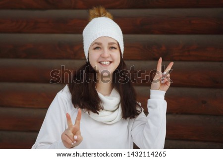 Young, pretty woman in a knitted white hat and sweater happy smiling on the background of a wooden brown log wall. Youth, healthy lifestyle, dreams.