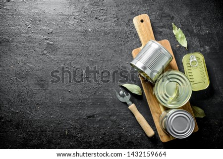 Closed cans of food on a wooden cutting Board with a opener and Bay leaf. On black rustic background