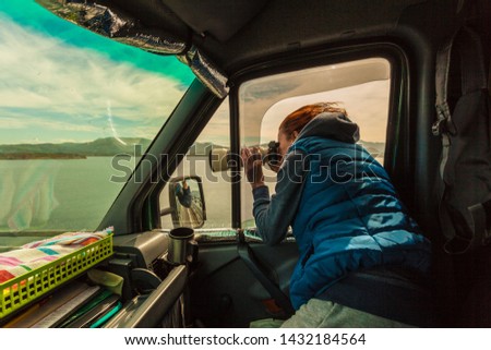 Mature woman tourist professional photographer taking photo from camper car with camera, driving on road trip. Female passenger taking picture out of window, windy weather