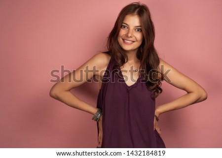 Young curly brunette puts her hands with wristlet on her hips smiling at the camera, pink background