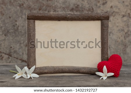 Brown wooden frame on the rough wooden table with the white millingonia and red knitting heart. Background of the rock wall. Sun light shines on the frame. Copy space for editing and text.