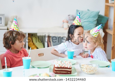 One of cute girls in birthday caps whispering something to her friend