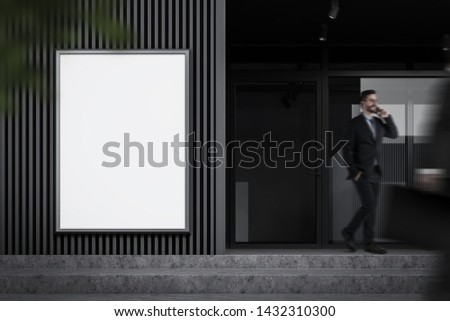 Young businessman on phone walking near office building with gray walls and large mock up poster for advertising. Concept of marketing. Blurred