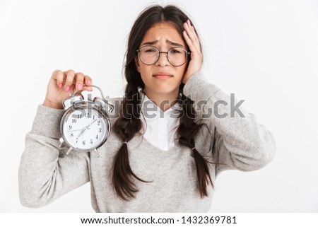 Photo closeup of concerned teenage girl wearing eyeglasses frowning while holding alarm clock in hands isolated over white background