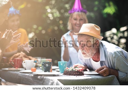 Senior birthday in. backyard tropical garden. White, asian senior man and woman with young couple. Enjoying a birthday party at home garden. Try to blow candle with light leaks. Royalty-Free Stock Photo #1432409078