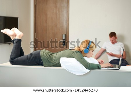 Pensive young woman in headphones programming on laptop whne her boyfriend texting friend in background