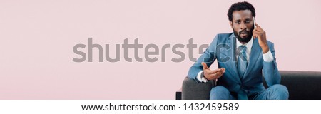 young african american businessman sitting in armchair and talking on smartphone, isolated on pink