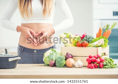 Woman with sports figure on her belly shows heart shape in home kitchen with wooden box full of organic vegetable.