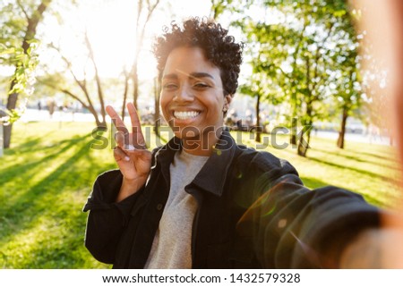 Photo of charming african american woman with curly hair showing peace sign and taking selfie on cellphone while walking in city park