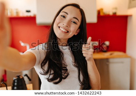 Long-haired woman dressed in white T-shirt makes selfie in kitchen and shows thumb up Royalty-Free Stock Photo #1432622243