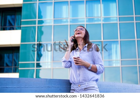 Happy smiling business woman talking on mobile smartphone and drinking coffee from disposable paper cup in the street with office buildings in the background.