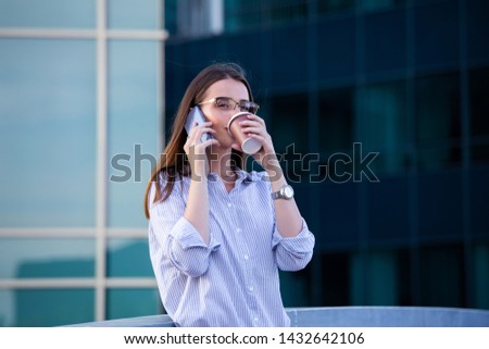 Executive business woman talking on mobile smartphone and drinking coffee from disposable paper cup in the street with office buildings in the background.