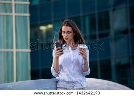 Executive business woman looking at mobile smartphone and drinking coffee from disposable paper cup in the street with office buildings in the background.