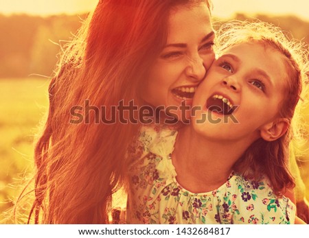 Happy enjoying mother hugging and biting her playful laughing kid girl on sunset bright summer background. Closeup toned portrait