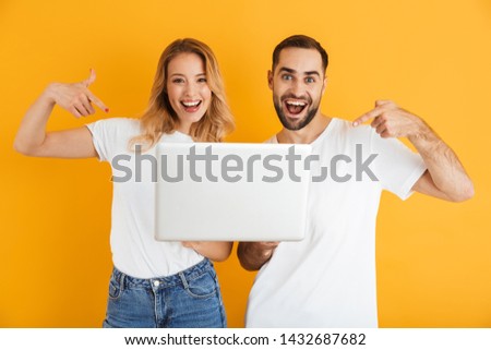 Image of excited couple man and woman rejoicing together while pointing fingers at laptop isolated over yellow background