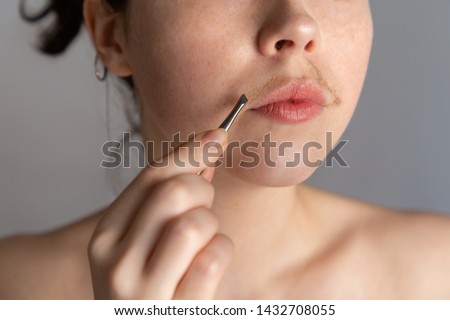 A young woman with a mustache tries to remove the hair over her lip with tweezers. The concept of getting rid of unwanted facial hair. Close up