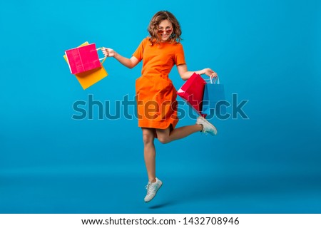 attractive happy smiling stylish woman shopaholic in orange trendy oversize dress jumping running holding shopping bags on blue studio background isolated, colorful, sale excited, summer fashion trend