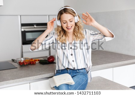 Image of a happy young woman posing at the kitchen at home listening music with headphones laughing writing notes.