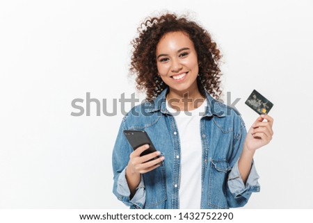 Portrait of a pretty cheerful casual african girl standing isolated over white background, showing plastic credit card, using mobile phone