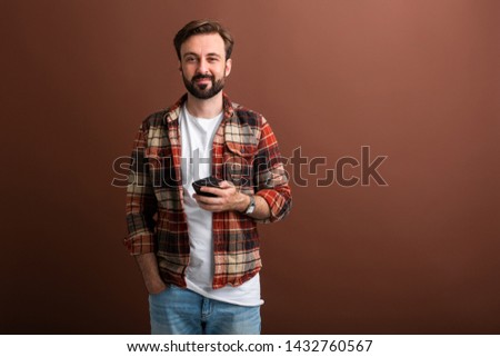 portrait of handsome stylish bearded man on brown background holding smart phone smiling isolated in vintage checkered shirt, looking in camera