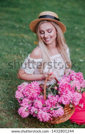 Beautiful girl in vintage dress and straw hat sitting and holding pink peonies in rustic basket in park