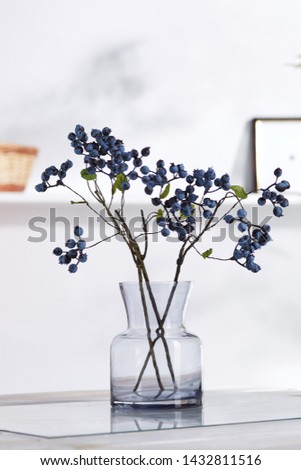 A closeup shot of an interior design. In the middle there is a composition with several blueberry twigs in a transparent glass vase. On the blurred background there is a wall shelf with photo frame.
