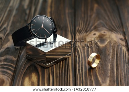 Black modern wrist watch on chrome cube with golden ring on wooden desk