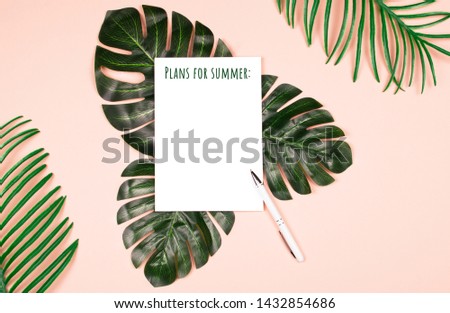 Beautiful shiny artificial leaves of jungle plants on pale pink background and note pad with white pen for writing. Elegant decoration elements with copy space for text. Tropical concept.