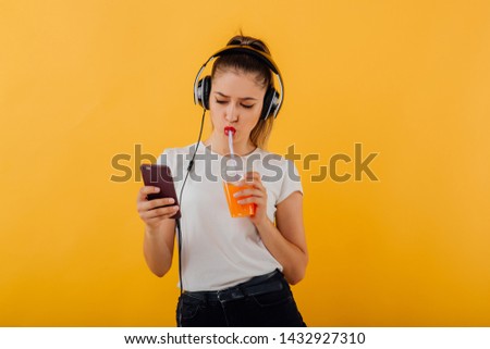 beautiful teenager, listening to music with headphones on head, looking in the phone, drink juice plastic cup, dressed in a white shirt,  isolated on a yellow background, copy space