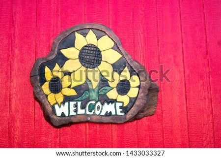 Wooden welcome sign featuring sunflowers is hung on a bright red wooden wall.
