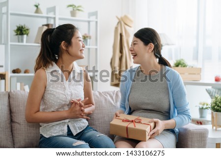 young best female friend visit bestie giving gift box cardboard to pregnant woman sitting on sofa couch. Happy mother in living room at home. Pregnancy and motherhood expect baby friendship concept.