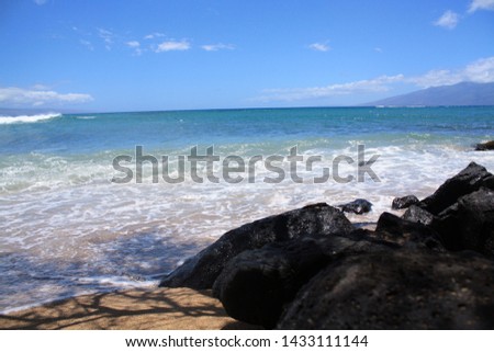 waves and lava rocks crash blue waters.  Mountains in background with  clear sky on a Maui, Hawaii beachfront.