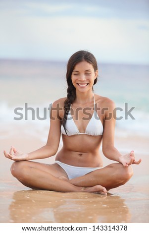 Meditation - woman on beach meditating by ocean sea smiling serene and happy. Asian girl sitting relaxing enjoying summer beach holidays travel. Multiracial Asian / Caucasian bikini model.