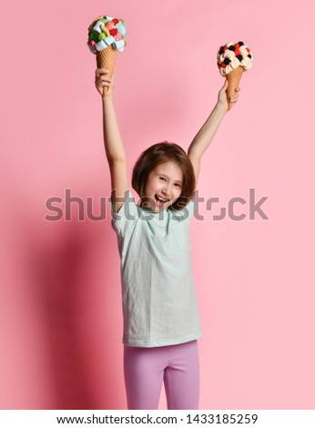 Happy shouting teen girl lifts up two big ice cream in waffles cone with raspberry marshmallow marmalade like she has won it on pink background