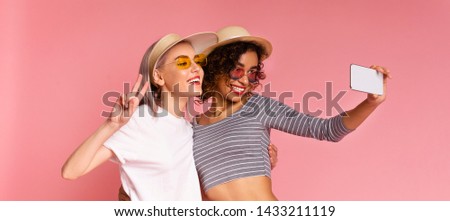 Two millennial girls taking selfie together, embracing and smiling at cellphone camera, pink studio background