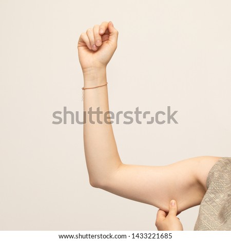 A closeup view of a young Caucasian lady squeezing her upper arm, showing the sagging skin and fat beneath the triceps, commonly referred to as bingo wings. Isolated against a white background. Royalty-Free Stock Photo #1433221685