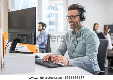 Smiling handsome male customer support phone operator with headset working in call centre. Group of sales agent working in office. Royalty-Free Stock Photo #1433366867